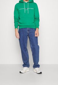Sudadera de algodón verde con bolsillo frontal, presenta texto de logo blanco; combinada con jeans de denim azul y zapatillas deportivas ligeras.