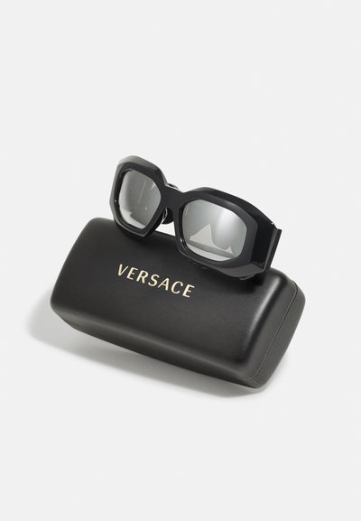 Mustat ylisuurteiset aurinkolasit harmailla linsseillä, joissa on paksu geometrinen kehys, lepäävät mustassa kotelossa, jonka kannessa lukee "VERSACE" kultakirjaimin.
