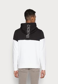 Mann von hinten, der einen schwarz-weißen Hoodie mit dem Text "CALVIN KLEIN" auf der Kapuze trägt, kombiniert mit hellgrauen Hosen.