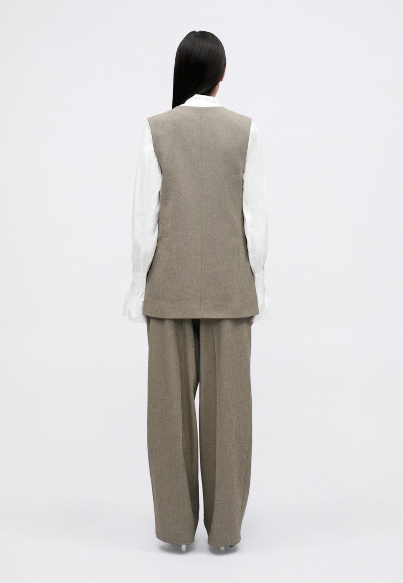 Tweed zonder mouwen vest in lichtbruin, gecombineerd met een losse beige broek. Witte blouse met lange mouwen en gerimpelde manchetten eronder.