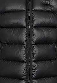 Sort pufferjakke med quiltede sektioner, lynlås foran og justerbare detaljer, lavet af et glat, letvægtsstof.