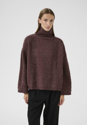 Femme portant un pull en col roulé maroon ample avec des panneaux verticaux texturés et un pantalon noir à jambe large, se tenant devant un fond blanc uni.