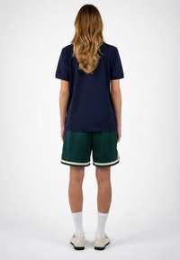 Femme aux longs cheveux ondulés, vue de dos, portant un t-shirt bleu marine, un short vert avec un bord blanc, des chaussettes blanches et des chaussures beiges.