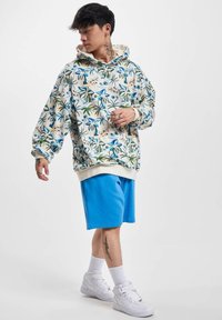 La sudadera con capucha presenta una base crema con patrones florales en azul y verde, combinada con unos pantalones cortos azules. Se usan zapatillas blancas y calcetines.
