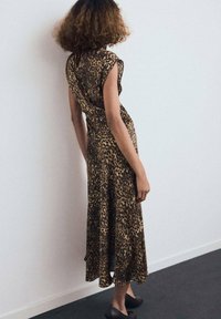 Mujer con cabello rizado, vestida con un vestido midi sin mangas de estampado de leopardo y zapatos negros, de pie contra una pared blanca sobre un suelo oscuro.