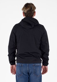 Giacca nera con cappuccio, polsini elasticizzati, materiale leggero e vestibilità comoda. Abbinata a jeans blu e vista da dietro.