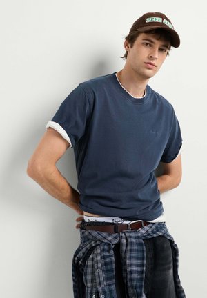Jonge man met een marineblauw T-shirt, een bruine pet, een geruit overhemd om de taille gebonden en een donkere broek, staand tegen een witte muur met handen achter de rug.