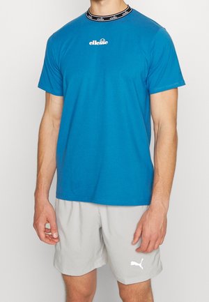 Mężczyzna w niebieskim t-shircie ellesse i jasnoszarych szortach Puma, stojący z ramionami swobodnie opuszczonymi wzdłuż ciała na jednolitym tle.