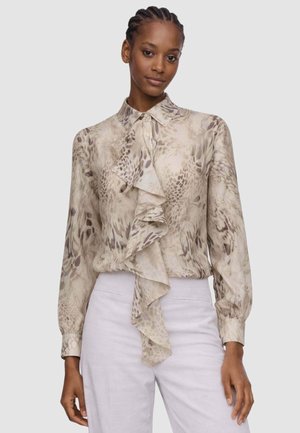 Donna che indossa una blusa beige con stampa animalier e una grande ruches frontale, abbinata a pantaloni chiari a vita alta, in piedi davanti a uno sfondo semplice.