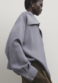 Chemise oversized à rayures bleues et blanches avec un grand col, un tissu texturé et des poignets arrondis ornés de boutons de couleur dorée.