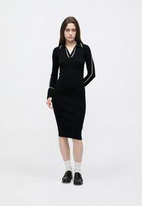 Μαύρο πλεκτό φόρεμα με μακριά μανίκια, V-λαιμόκοψη και λευκές λεπτομέρειες σε γιακά και μανίκια. Εφαρμοστή σιλουέτα, μήκος ως το γόνατο, λεία υφή.