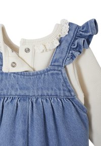 Denim-Overall mit einem hellblauen Waschung, ausgestattet mit einem rüschierten Schulterträger und einem weißen, gerippten Langarmshirt mit Spitzenbesatz am Ausschnitt.
