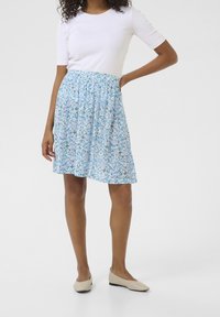 Blauer Blumenrock mit elastischem Bund, knielang, aus leichtem Material und mit geraffter Textur. Kombiniert mit einem weißen, taillierten Oberteil und beigen Ballerinas.