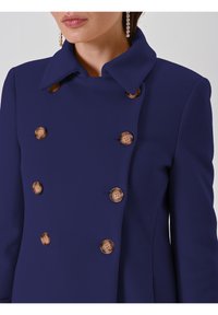 Cappotto doppiopetto blu navy con tessuto strutturato, ampio colletto e bottoni in legno. Vista ravvicinata che mette in evidenza i dettagli dei bottoni.