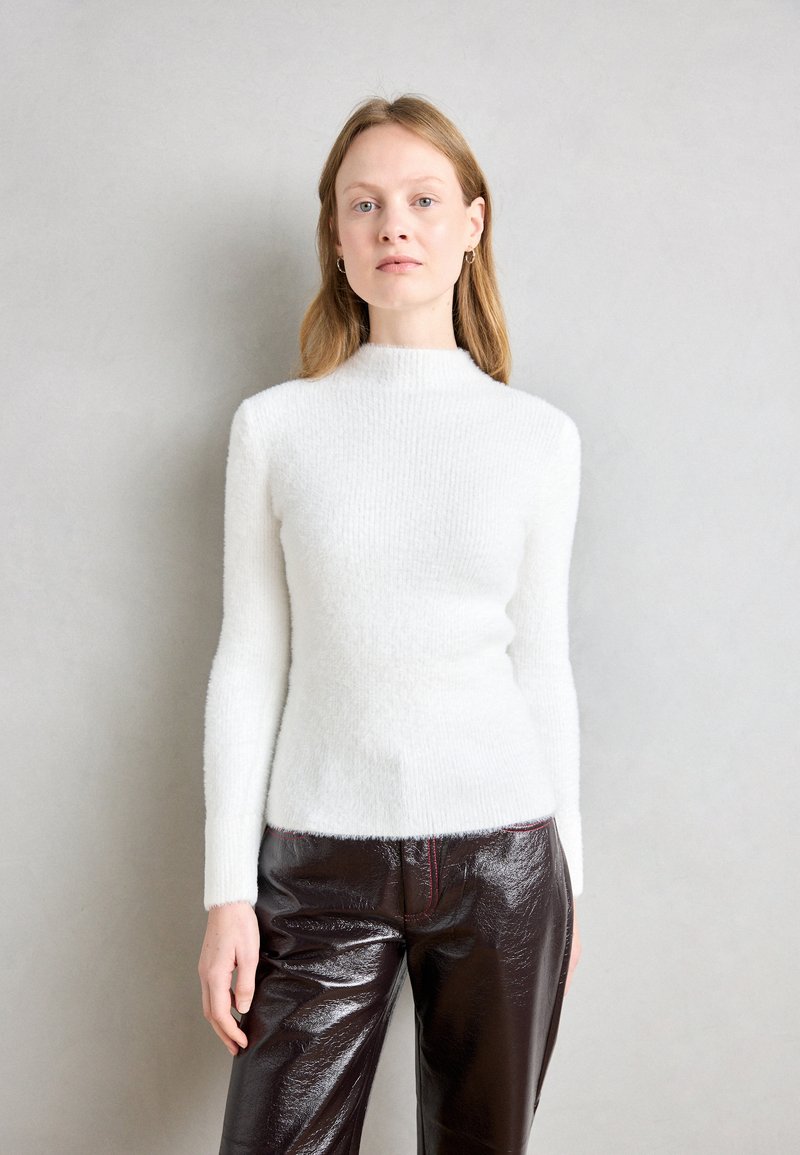 Weißer, flauschiger Strickpullover mit hohem Kragen und langen Ärmeln, kombiniert mit glänzenden, dunkelbraunen Lederhosen mit roter Naht.