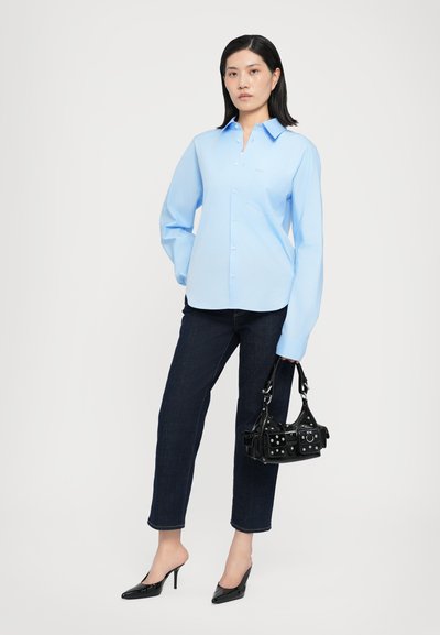 Light blue button-up shirt with a collar, long sleeves, and a single chest pocket paired with dark jeans and black high-heeled shoes.