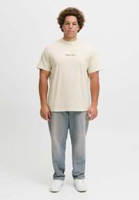T-shirt en coton beige à manches courtes et col rond. Présente un texte brodé en marron. Associé à un pantalon en denim bleu clair.