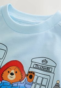 Svetlomodrá bavlnená mikina s ilustráciou medveďa v červenej čiapke a čiernymi obrysovými kresbami telefónnych búd.