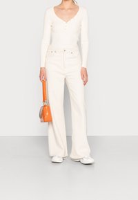 Kvinna iklädd en krämfärgad långärmad topp och vida byxor, som håller en liten orange handväska, står mot en enfärgad ljus bakgrund.