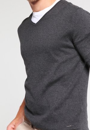 Mand iført mørkegrå V-hals sweater over hvid undertrøje, med beige bukser, stående mod en ensfarvet lys baggrund.