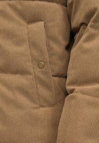 Bruin corduroy jasje met verticale ribbeltextuur, voorzien van een klepzak en een messing drukknoopdetail. Zacht, gewatteerd materiaal.