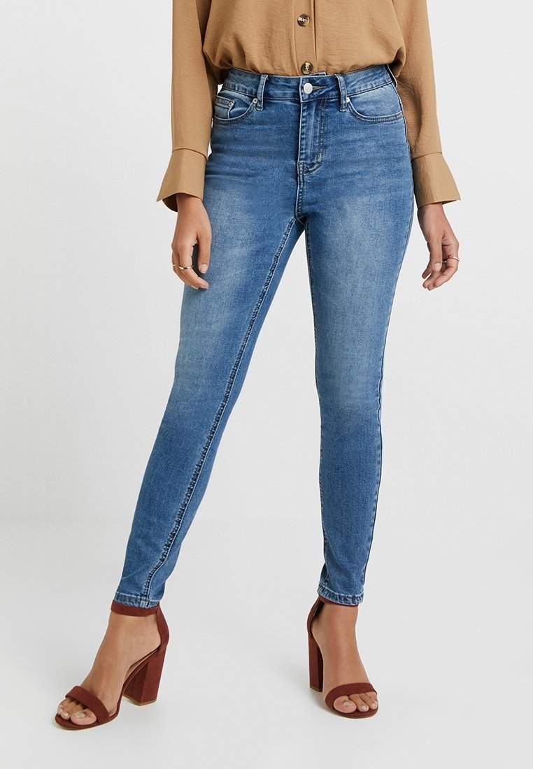 Jeans ajustados de denim azul claro, con una cintura alta, cinco bolsillos y un acabado suave. Llevados con una blusa marrón y tacones rojos de bloque.