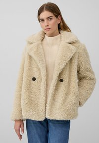 Jonge vrouw in een crèmekleurige, fluffy woljacke over een beige gebreide trui en blauwe jeans, staande tegen een effen achtergrond.