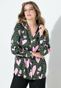 Groene blouse met opknopen met lichtroze hart en zwarte accentpatronen; heeft een kraag en lange mouwen en is gemaakt van een gladde stof.