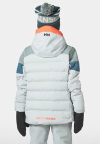 Bílá péřová bunda s modrými a šedými akcenty, vybavená kapucí a elastickými manžetami, doplněná černými rukavicemi a pruhovanou čepicí s bambulí.