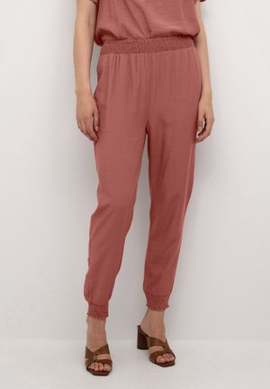 Pantalons roses amples avec taille smockée, poignets effilés et tissu léger. Associés à des sandales à talons carrés marron.