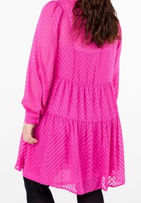 Růžové řasené šaty s dlouhými rukávy, vyrobené z texturované tkaniny s vzorem chevron. Průhledná vrstva a detaily na manžetách se sklady.