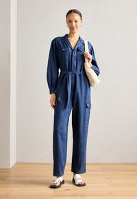 Jeansoverall i djupblått med långa ärmar, knytning vid midjan, fickor med lappar och raka ben. Bärs med vita sandaler och en liten beige väska.