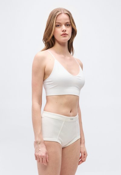 Νεαρή γυναίκα με λευκό κοντό τοπ και ψηλόμεσα σλιπ, στέκεται μπροστά σε απλό ανοιχτό γκρι φόντο.