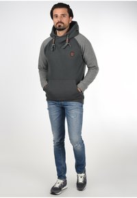 Sudadera con capucha en gris oscuro con mangas raglán grises, que cuenta con un bolsillo frontal y cordones. Combinada con jeans azules desgastados y zapatillas.