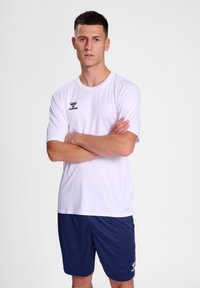 Hvit idretts T-skjorte med korte ermer og en liten svart logo; kombinert med marineblå shorts, glatt tekstur, og sporty passform.