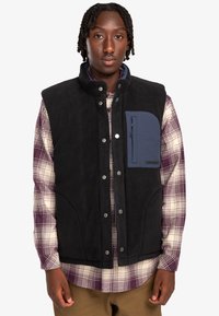 Black fleece vest with a snap closure, featuring a navy chest pocket. Worn over a plaid shirt with white, purple, and green hues.