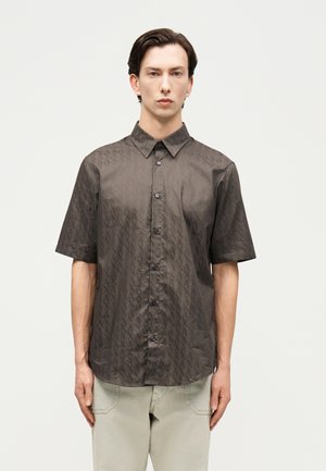 Mees, kes kannab tumepruuni lühikeste varrukatega nööpidega särki, millel on õrn teemantmuster, ning heledavärvilisi pükse, taust on lihtne.