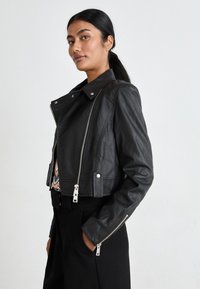 Veste en cuir noir croppée avec col à revers et quincaillerie argentée, dotée de fermetures éclair à l'avant et de poignets accentués. Texture lisse.