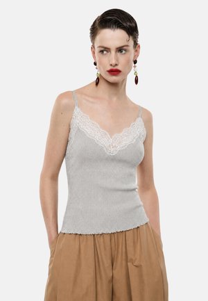 Donna con top grigio a coste senza maniche con bordo in pizzo bianco e pantaloni marroni larghi, che guarda di lato con le mani in tasca.
