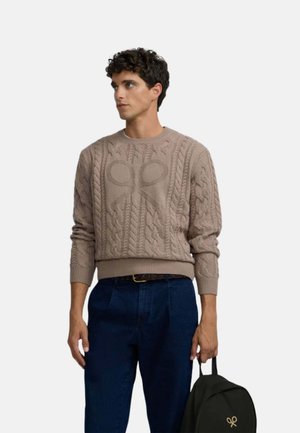 Hombre con suéter beige de punto trenzado y pantalones azul oscuro, sosteniendo una mochila negra con un logo dorado en una mano.