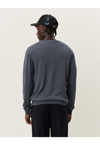 Grauer Pullover mit langen Ärmeln und rundem Halsausschnitt, aus strukturiertem Stoff. Getragen mit dunklen Hosen. Eine schwarze Mütze mit bunten Stickereien.