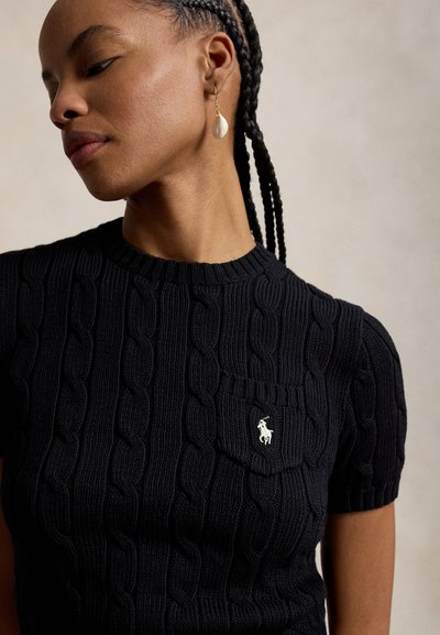 Mujer con el cabello trenzado, que lleva un suéter de manga corta de punto de cable negro con un pequeño logo bordado en el bolsillo del pecho y un pendiente en forma de concha.