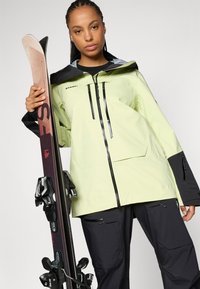 Giacca impermeabile verde chiaro con cappuccio e dettagli neri; presenta una chiusura con zip e tasche laterali. Tiene uno sci con attacchi.