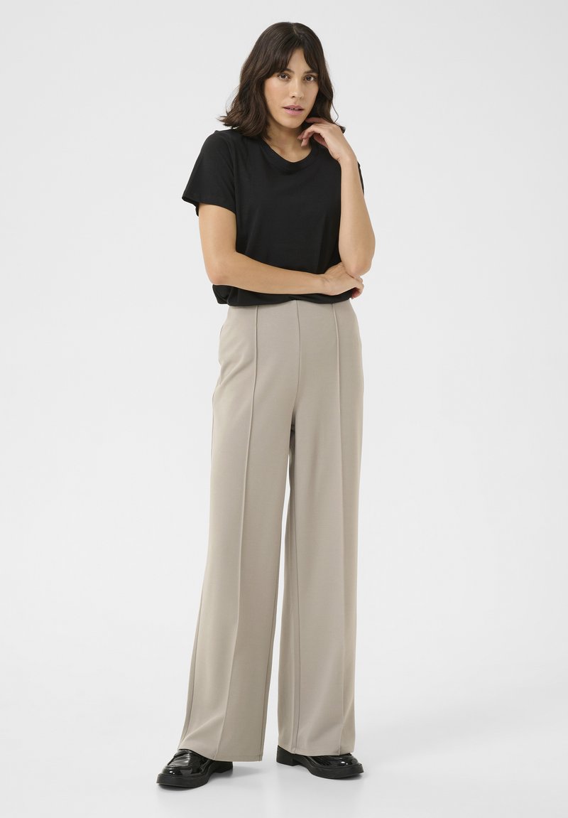 Zwart t-shirt met korte mouwen, gecombineerd met beige wijde broek met verticale naden, en zwarte glanzende schoenen, tegen een effen achtergrond.