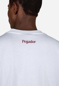 Valge T-särk ümarate kaelustega ja lühikeste varrukatega. Ülaosas seljal on marooni värvi "Pegador" tekstilogoga. Pehme kangast tekstuur.