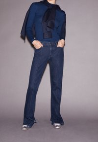 Jean évasé bleu foncé avec surpiqûres blanches contrastantes, associé à un haut côtelé bleu marine et une écharpe nouée en guise d'accent. Tissu lisse et coupe structur ée.