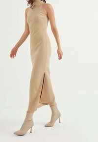 Beige ribbed maxi dress with a high neckline and side slit, paired with beige pointed-toe ankle boots, showing a smooth texture and fitted design.