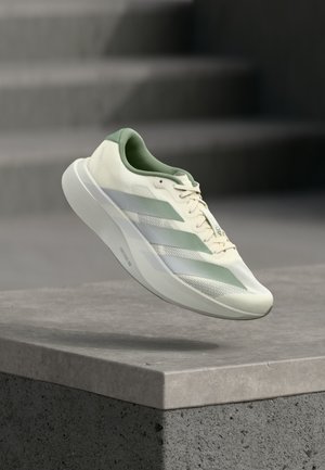 Witte en groene sportieve sneaker met drie strepen zweeft boven een betonnen richel en trappen in zacht licht.