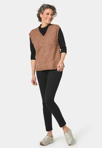 Bruine, mouwloze breivest met een V-hals, gedragen over een zwart langmodieuze top. Gemaakt in combinatie met zwarte slim-fit broek en lichtbeige sneakers.