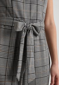 Šedá a černá kostkovaná látka s vázací stuhou. Materiál má strukturovaný povrch s různými vzory a zdobným prošíváním.